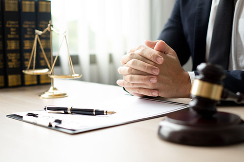 Hands clasped over a document on a desk with a scale of justice and a wooden gavel nearby.